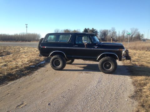 1979 Ford Bronco - A lifted black 1979 Ford Bronco is parked on a gravel road with dry grass and trees in the background under a clear blue sky.