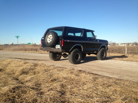 1979 Ford Bronco - A black 1979 Ford Bronco is parked on a dirt road with dry grass and a harvested field in the background. The rear-mounted spare tire is vi…