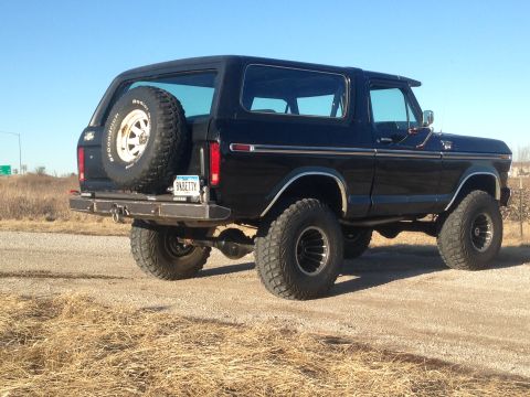 1979 Ford Bronco - The rear driver's side of a black 1979 Ford Bronco is shown parked on a gravel road with dry grass in the foreground. A spare tire is mount…