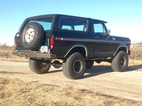 1979 Ford Bronco - A black 1979 Ford Bronco is parked on a dirt road with dry grass on the side. The rear of the vehicle is visible, featuring a spare tire mo…