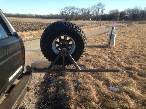 1979 Ford Bronco - A large spare tire is mounted on a custom metal rack attached to the rear of a dark-colored SUV. The vehicle is parked on a dirt road next…