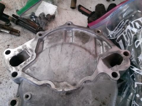 1996 Ford Bronco - A close-up, overhead view of a used, aluminum water pump housing with two rectangular openings and several bolt holes. The housing is resti…