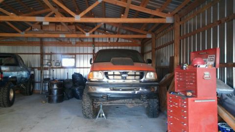 1982 Ford Bronco - An orange Ford F-150 pickup truck with a hood scoop is parked inside a metal-sided garage. A blue pickup truck is parked to the left of the…