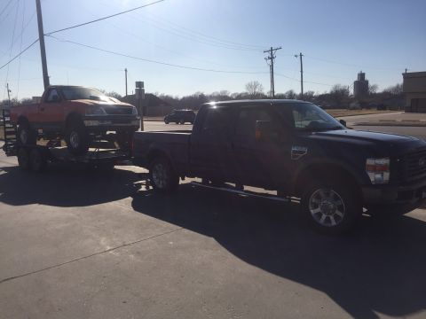 1982 Ford Bronco - A dark blue Ford F-250 Super Duty pickup truck tows a trailer carrying an orange Ford pickup truck. The scene appears to be on a sunny day…