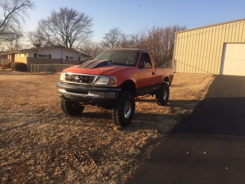 1982 Ford Bronco - A lifted, orange pickup truck with a black hood scoop is parked on a dry, grassy lawn in front of a house and a metal building. The truck h…