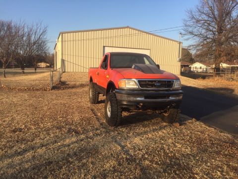 1982 Ford Bronco - A lifted, orange Ford F-150 pickup truck with a black hood scoop is parked on a dry, grassy area in front of a tan metal building. The truc…