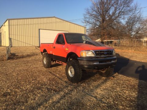 1982 Ford Bronco - A lifted, bright orange Ford F-150 pickup truck with a black hood scoop is parked in a grassy yard in front of a beige metal building.