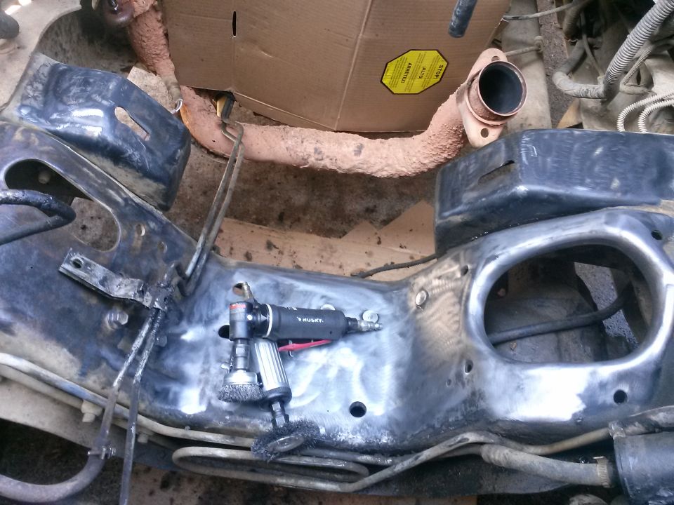 Vehicle frame being cleaned with a grinder, rusty exhaust pipe in background.