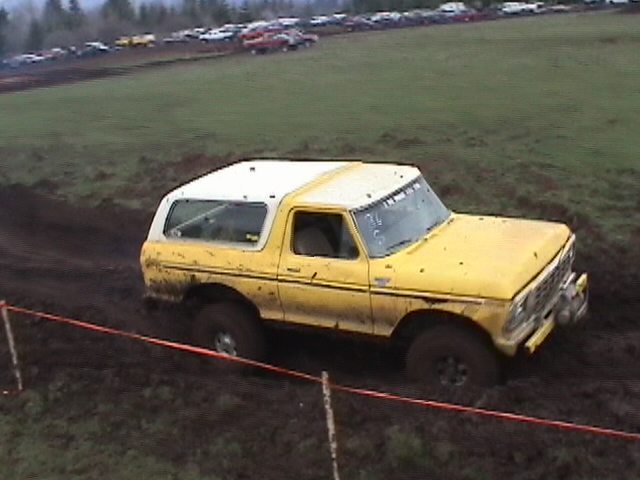 Yellow Ford Bronco with white top running a muddy off-road course at an event
