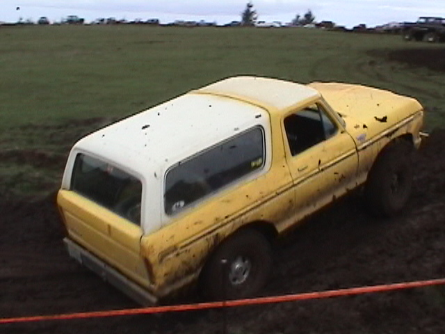 Muddy yellow Ford Bronco with white hardtop sitting in a dirt mud bog pit
