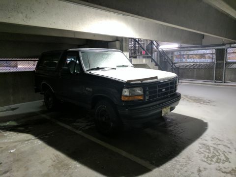 1995 Ford Bronco - A black 1995 Ford Bronco is parked in a concrete parking garage with snow on the ground and on the vehicle's hood. The front of the SUV is…