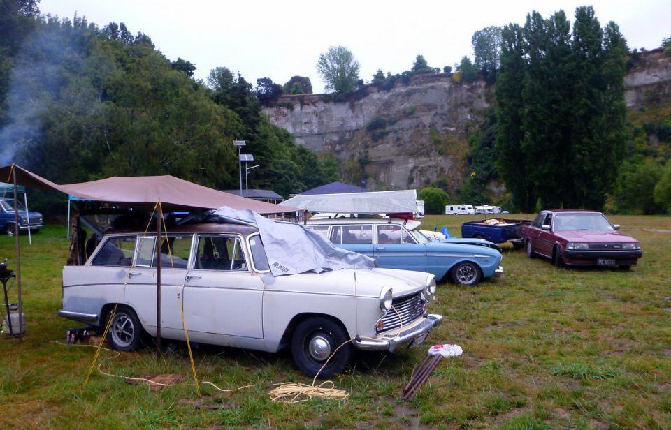 White vintage station wagon parked under a tarp, with other cars and a cliff in the background.