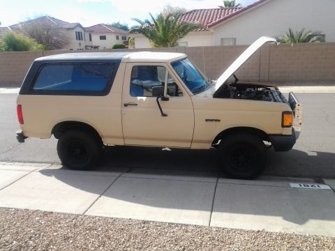 1991 Ford Bronco - A tan Ford Bronco with its hood up sits parked on a sidewalk in front of a residential building.