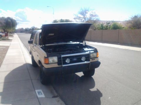 1991 Ford Bronco - A tan 1991 Ford Bronco is parked on the side of a residential street with its hood open, revealing the engine bay.