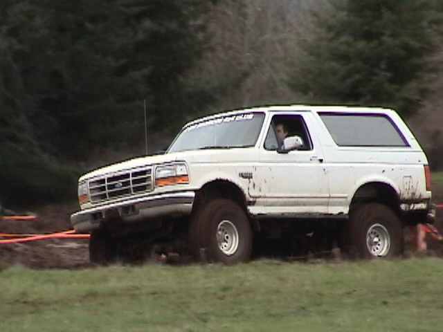 White Ford Bronco with windshield club lettering and driver inside crossing muddy grass field