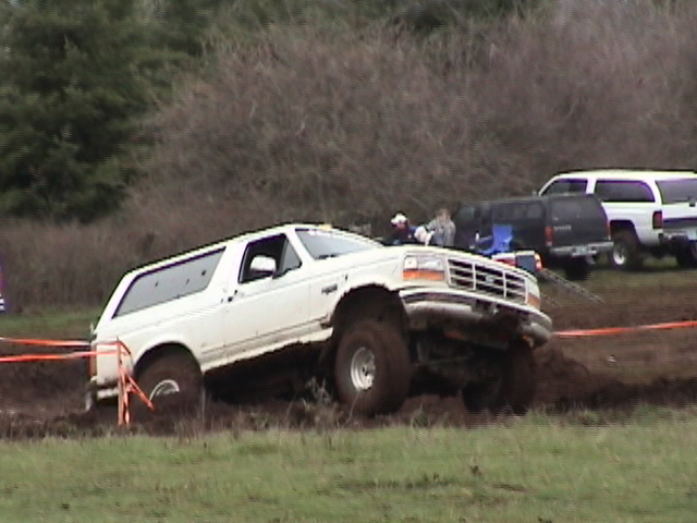 White Ford Bronco tilted in deep mud pit with front wheel raised at off-road event
