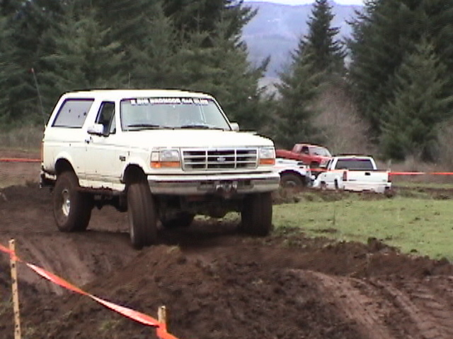 White Ford Bronco with club windshield text climbing muddy bank, evergreen trees behind