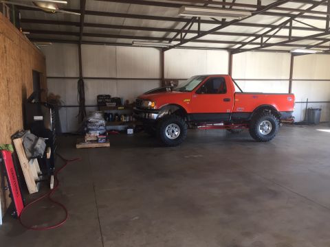 1982 Ford Bronco - A lifted, orange, two-door pickup truck with a black trim and large tires is parked inside a garage. Various tools and car parts are visibl…