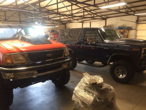 1982 Ford Bronco - Two modified trucks are parked side-by-side in a garage. The truck on the left is orange with a large hood scoop, and the truck on the righ…