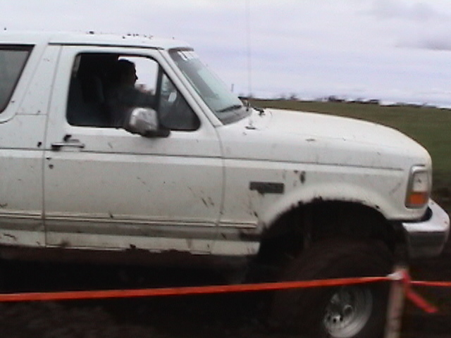 Close side view of muddy white Ford Bronco cab with driver inside on big tire in field