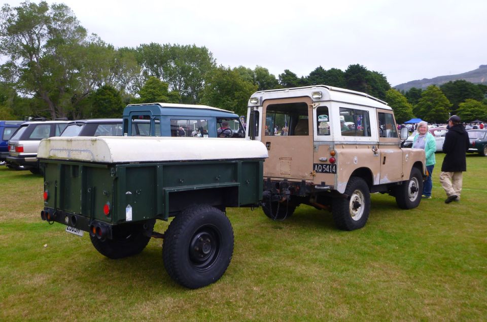 Tan and white Land Rover Series III parked next to a green trailer on grass.