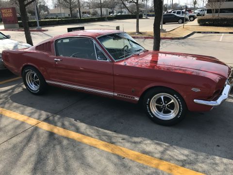 1965 Ford Mustang - A red 1965 Ford Mustang coupe is parked in a parking lot, viewed from the driver's side angle. The car features chrome trim, classic wheels…