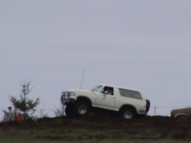 White lifted Ford Bronco on big tires cresting a dirt mound against gray sky, child watching