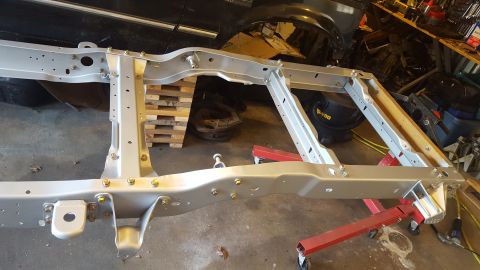 1994 Ford Bronco - A bare, silver-painted truck frame is supported by red jack stands in a workshop setting. Several bolts and brackets are visible along the…