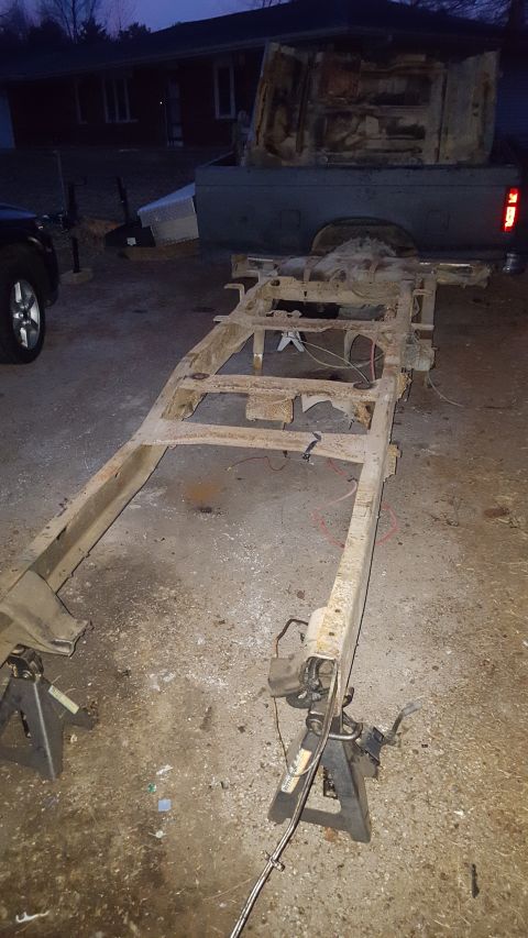 1994 Ford Bronco - A heavily rusted truck frame is supported by jack stands in a driveway at dusk. A portion of a pickup truck bed is visible in the backgroun…