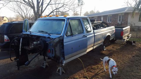 1994 Ford Bronco - A light blue pickup truck is parked outside a house, with its front end disassembled and supported by jack stands. A black SUV is parked be…