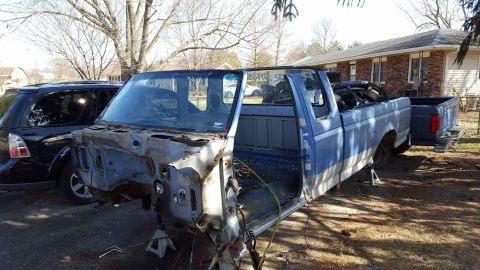 1994 Ford Bronco - A partially disassembled blue pickup truck is on jack stands in a yard with a house and trees in the background. A black SUV is parked to t…