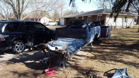 1994 Ford Bronco - A disassembled blue pickup truck chassis is supported by jack stands in a yard, with a black SUV parked to its left. Various vehicle parts…
