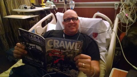 1985 Jeep CJ7 - A man in a hospital bed smiles while holding two magazines. One magazine has "YUKON RECOVERY GEAR" and the other has "CRAWL" with "MORE THA…