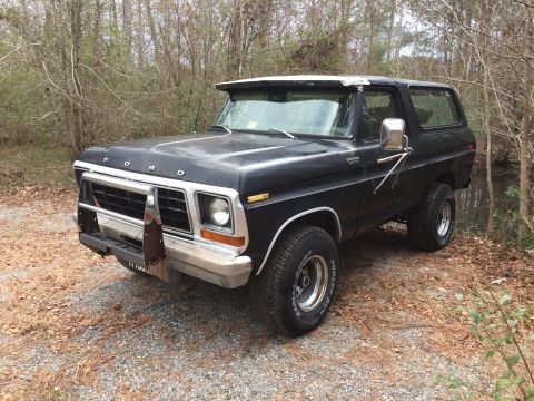 1979 Ford Bronco - A black 1979 Ford Bronco is parked on a gravel road surrounded by trees. The vehicle has a chrome grille, a custom front bumper, and a whit…