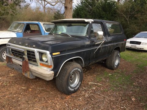 1979 Ford Bronco - A black 1979 Ford Bronco is parked outdoors on a dirt and grass lot next to a blue and white Ford truck. The Bronco has a chrome grille, a…