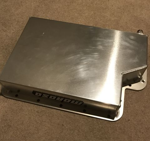 1995 Ford Bronco - A brushed aluminum Moroso oil pan sits on a carpeted floor. The oil pan has a visible logo and mounting holes along its edge.