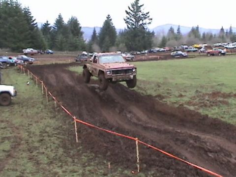 1994 Ford Explorer - A red pickup truck jumps over a muddy track at an off-road event, with spectators and other vehicles in the background.