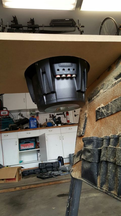 1996 Ford Bronco - A large black subwoofer is mounted upside down under a wooden shelf in what appears to be a garage or workshop. The background shows a work…