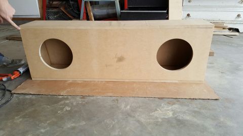 1996 Ford Bronco - A person's hand points to the corner of a large, unfinished wooden subwoofer box with two circular cutouts. The box sits on a piece of card…
