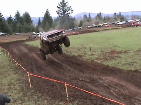1994 Ford Explorer - Off-road vehicle jumps a muddy track during a race event with spectators and other vehicles present.
