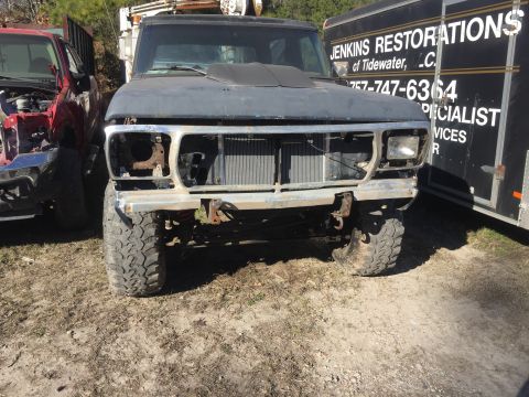 1979 Ford Bronco - The front of a dark blue, lifted Ford truck is shown, missing its grille and headlights. A black trailer with white lettering is parked to…