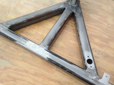 1979 Ford Bronco - A close-up, angled view of a metal triangular frame, likely a part of a vehicle chassis, resting on a wooden surface.