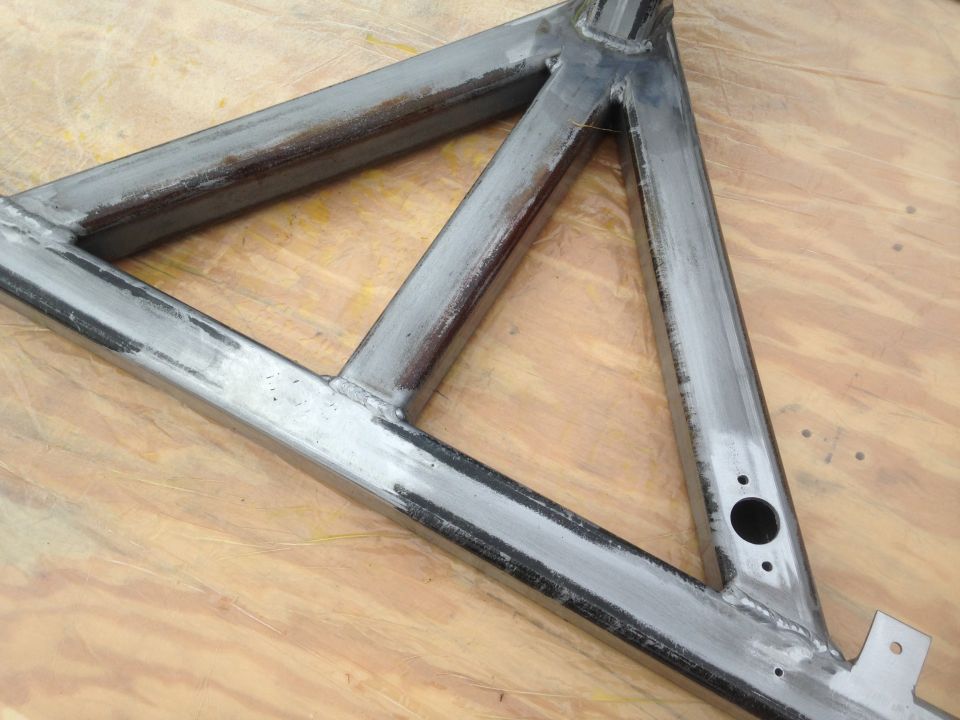 Metal triangular frame with visible welds and holes, resting on a wooden surface.