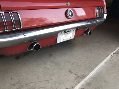 1965 Ford Mustang - The rear of a red 1965 Ford Mustang is shown, featuring dual exhaust pipes, a chrome bumper, and a Texas license plate.