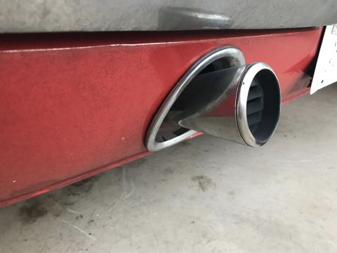 1965 Ford Mustang - Close-up of a chrome exhaust tip integrated into the red rear bumper of a vehicle. The exhaust tip has internal fins and is angled outwards.