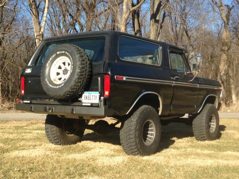 1979 Ford Bronco - The rear driver's side quarter view of a black 1979 Ford Bronco is shown parked on a grassy area with trees in the background. The vehicle…