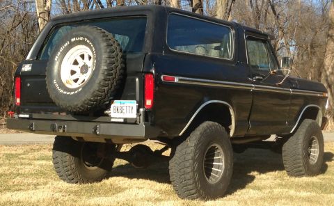 1979 Ford Bronco - The rear passenger side of a black Ford Bronco is shown parked on a grassy area with trees in the background. A spare tire is mounted on th…
