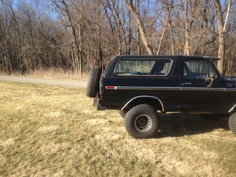 1979 Ford Bronco - The rear driver's side of a black 1979 Ford Bronco is shown parked on a grassy field next to a dirt road. A spare tire is mounted on the re…