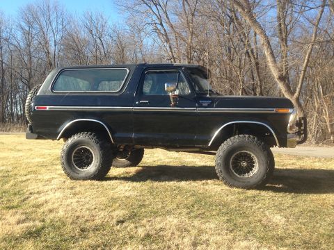 1979 Ford Bronco - The side view of a lifted black 1979 Ford Bronco is parked on a grassy field with bare trees in the background under a clear blue sky. The…