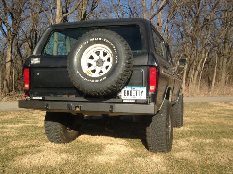 1979 Ford Bronco - The rear driver's side of a black 1979 Ford Bronco is shown parked on a grassy area with trees in the background. It features a spare tire…